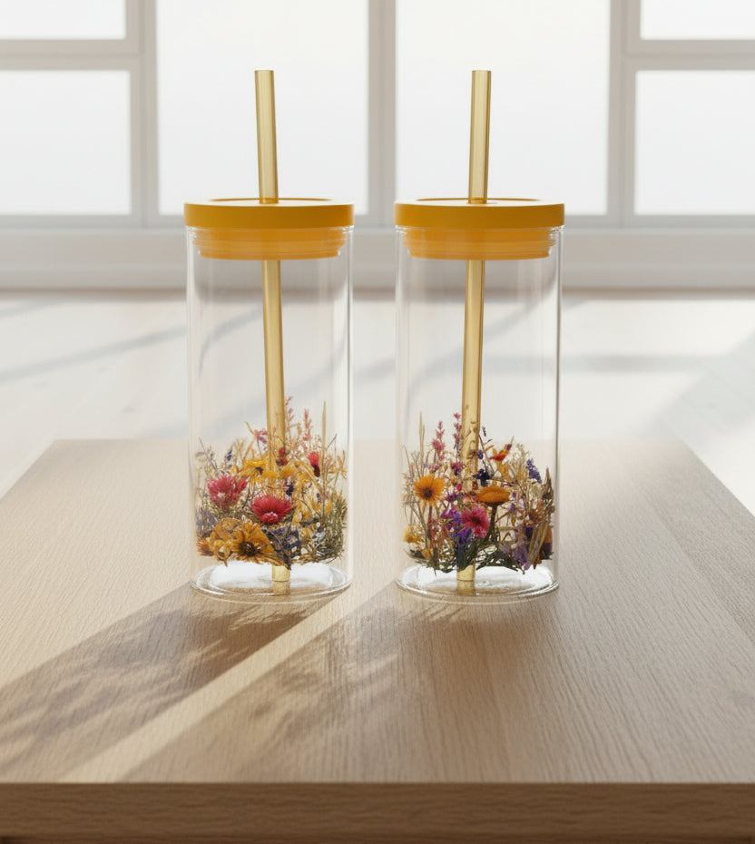 Two clear Glass cups with yellow lids and straws, decorated with colorful wildflower patterns near the bottom, set against a dark background.