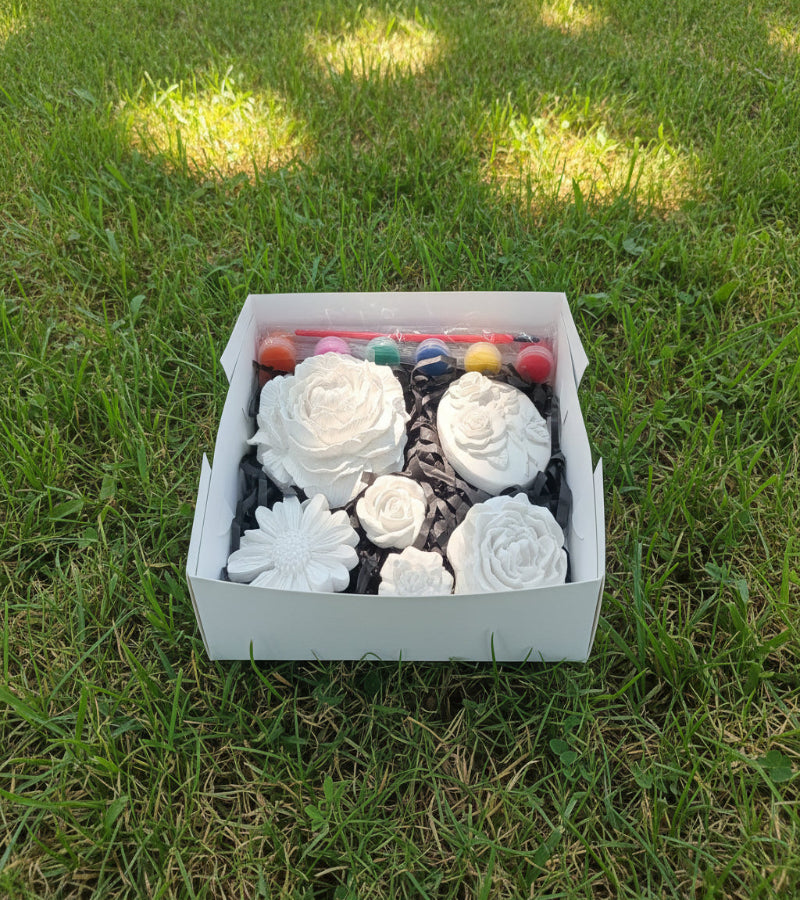 A white box containing Six unpainted white ceramic floral decorations (roses, daisy, and oval medallion) nestled in black shredded paper, with a set of colorful paint pots and painting brush.