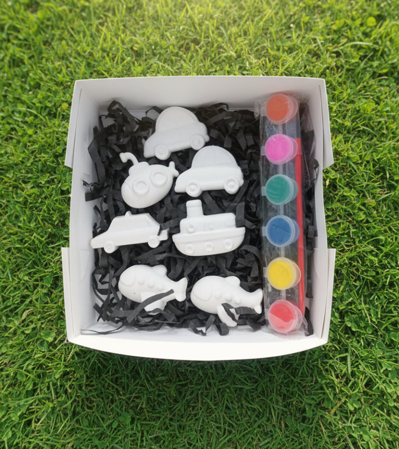 A box filled with black shredded paper holding seven unpainted white ceramic ship-shaped pieces and six colorful paint pots.