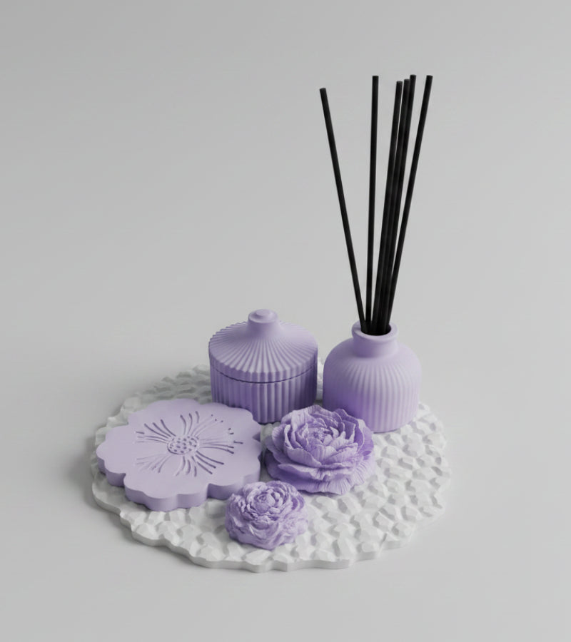 A lavender reed diffuser set on a textured white mat, featuring a small bottle with black reeds, a matching lidded jar, and two flower-shaped diffuser pieces