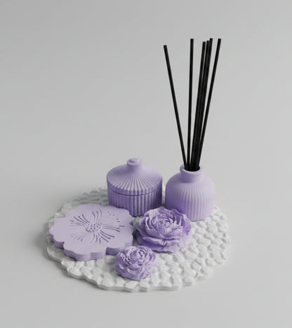A lavender reed diffuser set on a textured white mat, featuring a small bottle with black reeds, a matching lidded jar, and two flower-shaped diffuser pieces