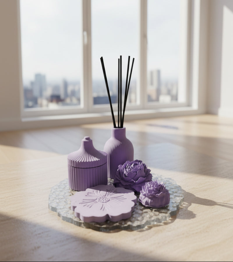 A set of lavender purple decorative items, including a flower-shaped coaster, two flowers, a small vase with reeds, and a small lidded container, arranged on a gray textured tray over a wooden surface.