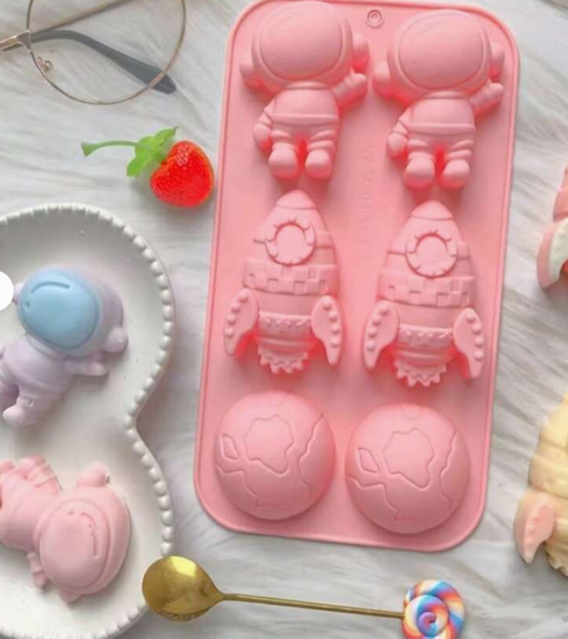 A pink silicone mold tray with four astronaut and rocket-shaped cavities, shown on a white textured surface with small decorative props including a strawberry, spoon, and pastel figurines.