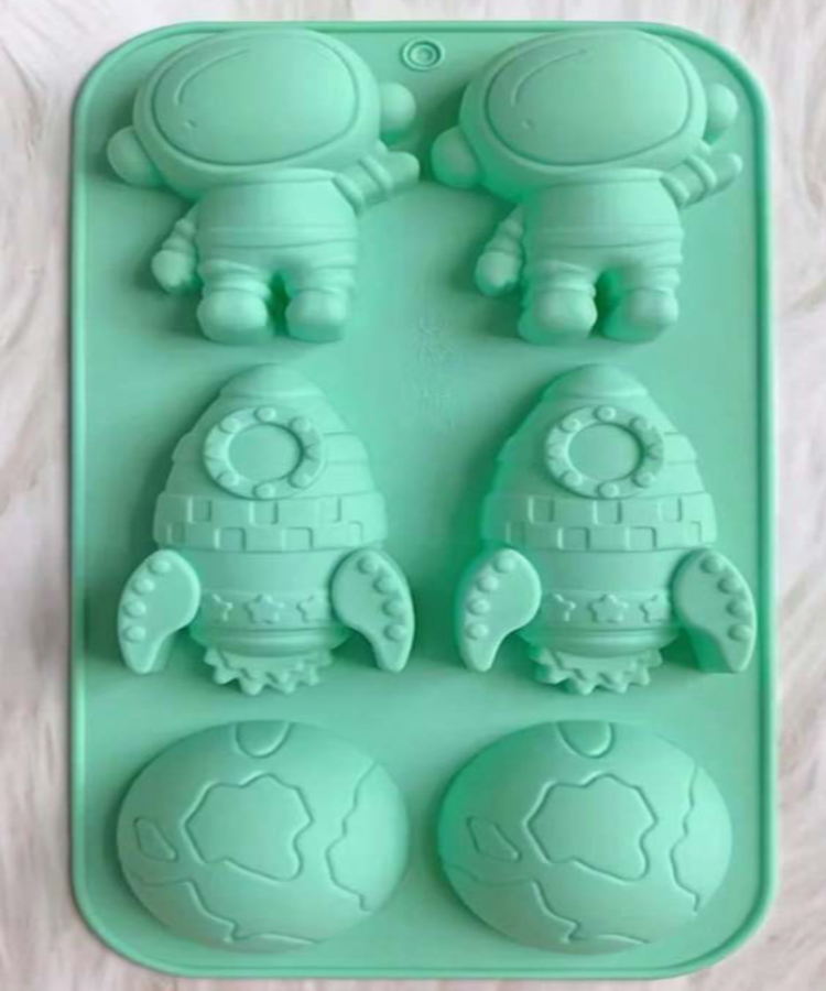 A mint green silicone mold tray featuring cartoon astronaut and rocket ship cavities, with two globe-shaped cavities on the right, shown on a light wood surface.