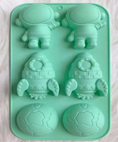 A mint green silicone mold tray featuring cartoon astronaut and rocket ship cavities, with two globe-shaped cavities on the right, shown on a light wood surface.
