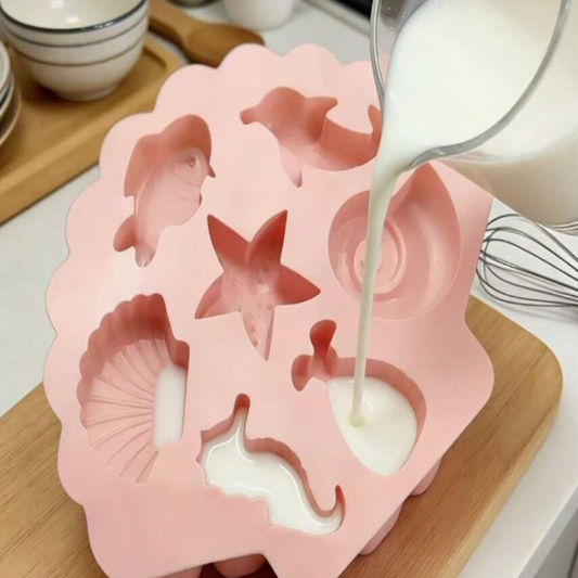 A pink silicone mold with sea-themed cavities including a starfish, seashell, dolphin, and wave, shown with white liquid being poured into one of the cavities on a wooden cutting board.