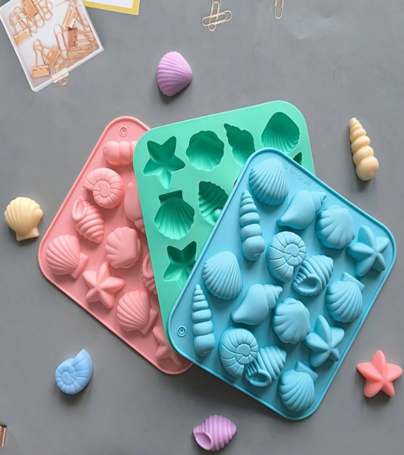 Two silicone mold trays with seashell and starfish cavities, one pink and one teal, shown on a gray surface with small seashell props around them.