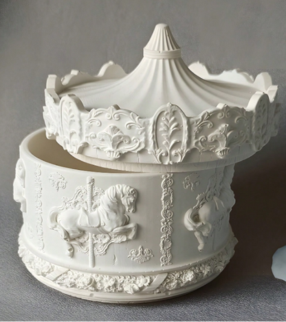 A white decorative ceramic or stone container with a matching lid, featuring embossed carousel horse and floral scrollwork details, photographed against a soft gray background.