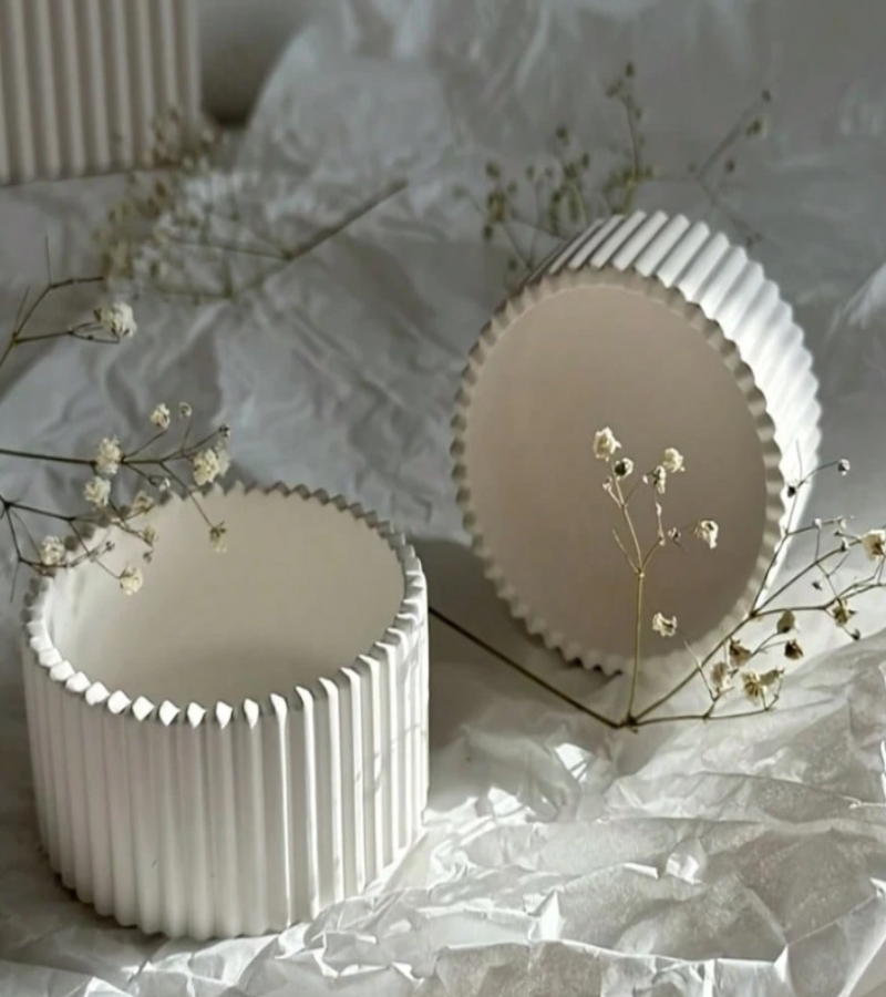 Two white ribbed ceramic storage or candle holders with scalloped rims, styled on crumpled white paper with small white flowers.