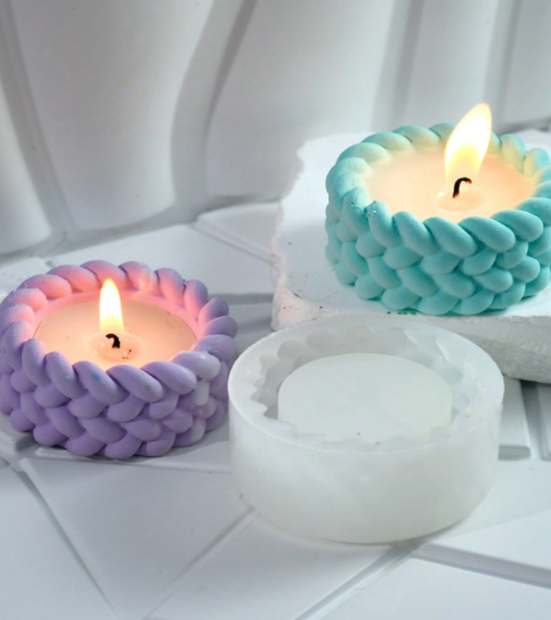 Three braided candle holders or candle cups shown on a white tiled surface, with two lit candles in pastel purple and aqua blue and one unlit white candle holder in front.