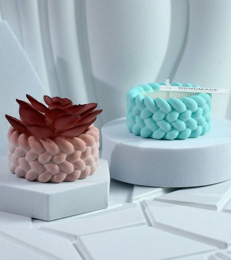 Two decorative candle products displayed on white pedestals in a clean studio setting. One candle has a pink textured base and a dark red succulent-shaped top.