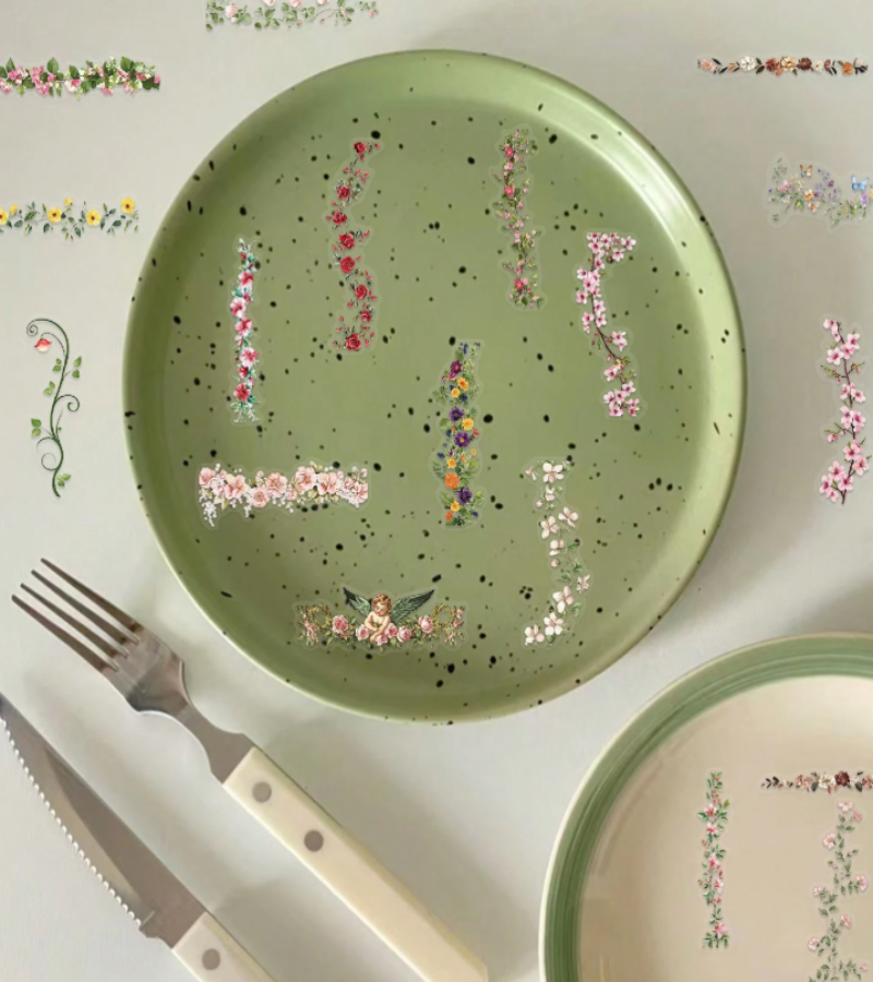 A top-down view of a light sage green ceramic plate decorated with PET floral Stickers and black speckles, styled with a fork and knife on a white surface with other floral plates around it.