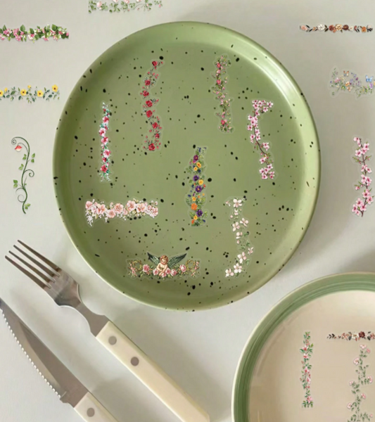 A top-down view of a light sage green ceramic plate decorated with PET floral Stickers and black speckles, styled with a fork and knife on a white surface with other floral plates around it.