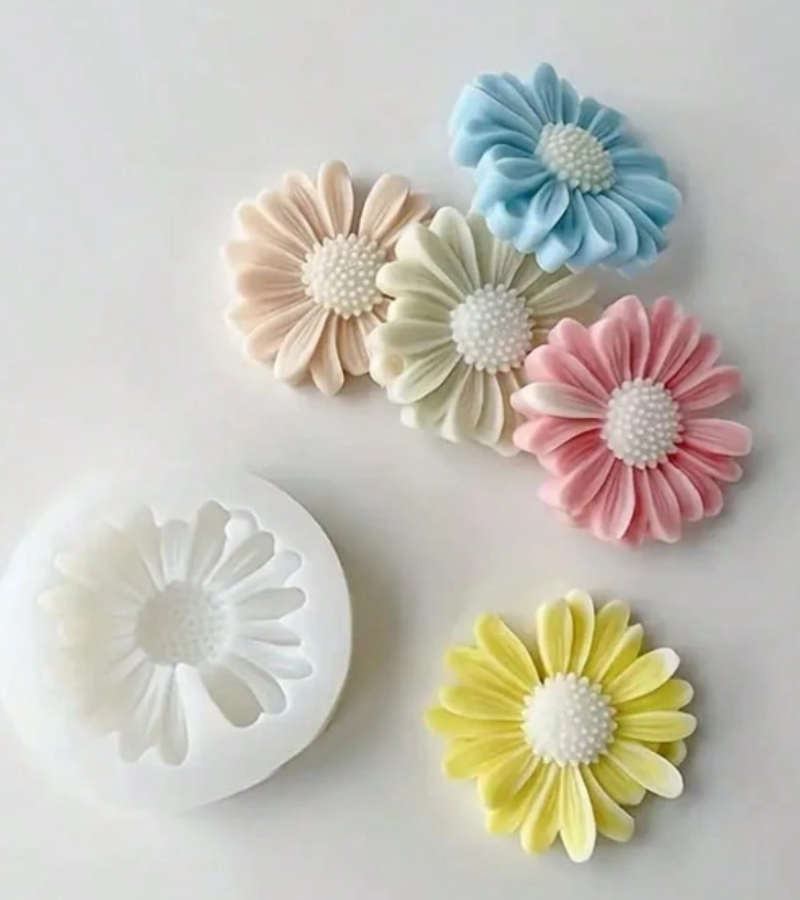 Five pastel daisy-shaped decorative pieces in cream, light blue, pale green, pink, and yellow, arranged on a white background beside a white mold.