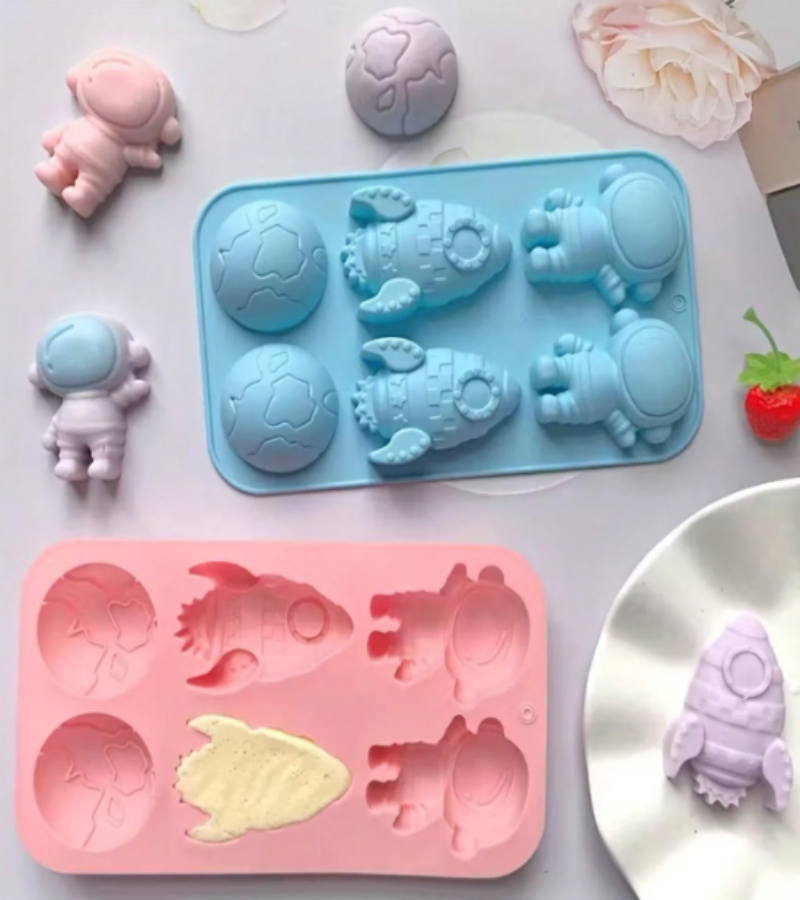 A set of pastel-colored astronaut-themed molds, including a light blue rectangular mold with six astronaut-shaped cavities, a pink rectangular mold with six cavities, and several small molded astronaut figurines in pink, lavender, and white arranged around them on a white surface.