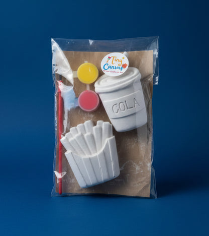 A miniature paint kit in a clear plastic bag, featuring a white cola cup labeled 'COLA', french fries, yellow and pink paint pots, and a paintbrush on a brown card.
