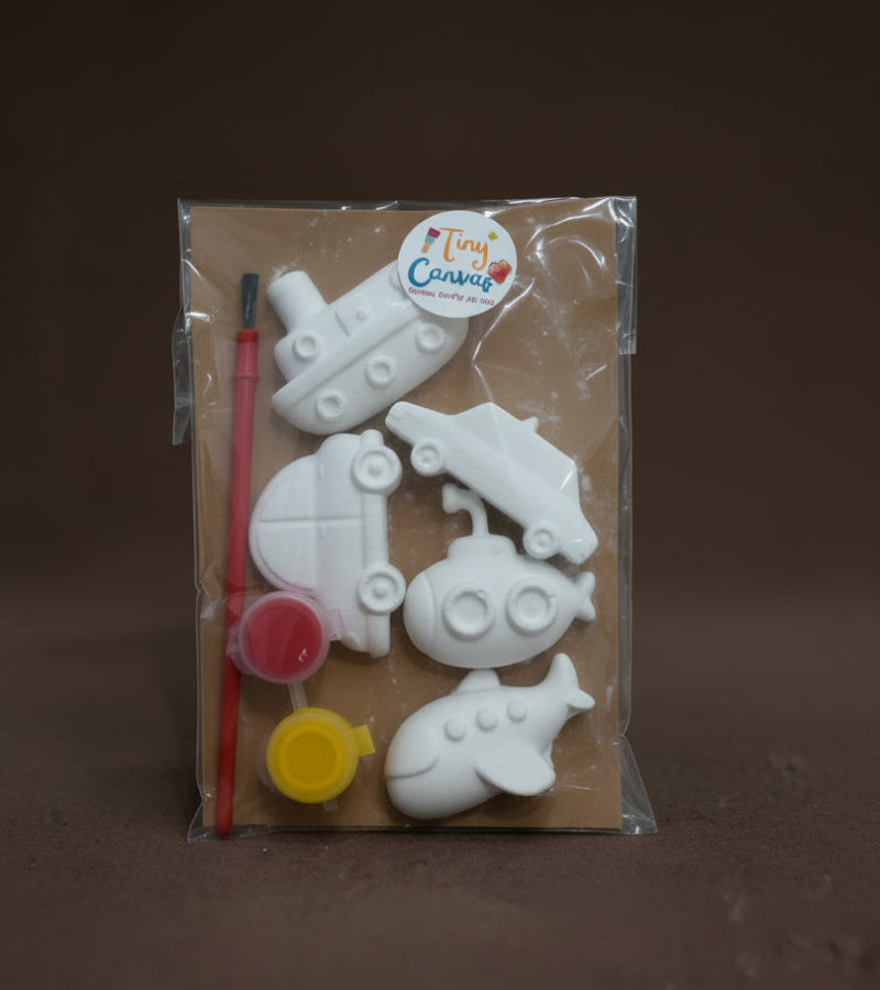 A children's DIY craft kit in clear plastic packaging, featuring transport theme, white unpainted car and ship, a paintbrush, and small paint pots on a brown card.