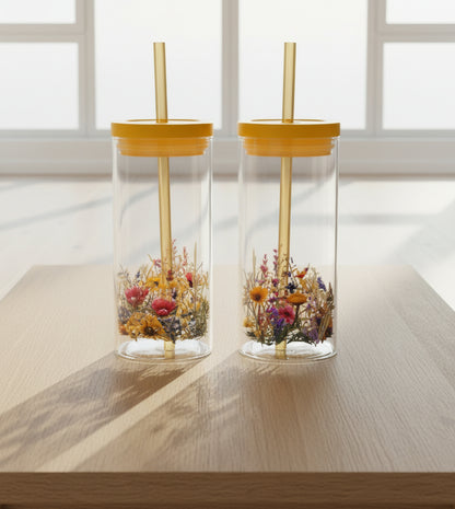 Two clear Glass cups with yellow lids and straws, decorated with colorful wildflower patterns near the bottom, set against a dark background.