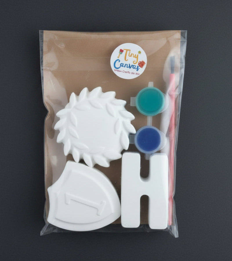 A packaged craft kit containing a white letter 'H', a white shield-shaped piece, laurel wreath piece, two small paint pots (green and blue), and a red-handled brush, all inside a clear plastic bag with a 'Tiny Canvas' logo.