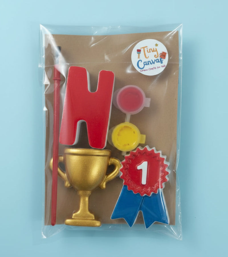 A packaged craft kit by Tiny Canvas containing letters and shapes, a small trophy, paint pots, and a brush, sealed in a clear plastic bag on a brown background.