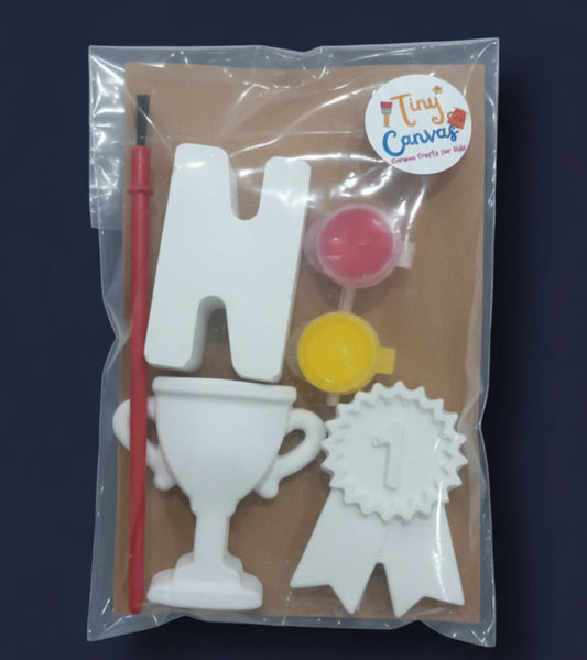 A packaged craft kit by Tiny Canvas containing white letters and shapes, a small trophy, paint pots, and a brush, sealed in a clear plastic bag on a brown background.
