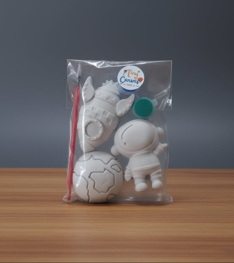 A clear plastic bag containing unpainted white figurines for kids, including a planet, a rocket ship, an astronaut, a paintbrush, with two small round paint container labeled 'Tiny Canvas'.