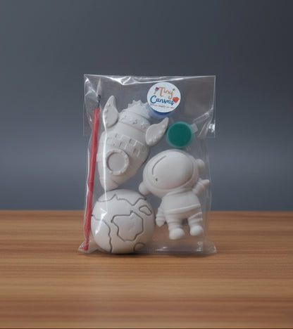 A clear plastic bag containing unpainted white figurines for kids, including a planet, a rocket ship, an astronaut, a paintbrush, with two small round paint container labeled 'Tiny Canvas'.