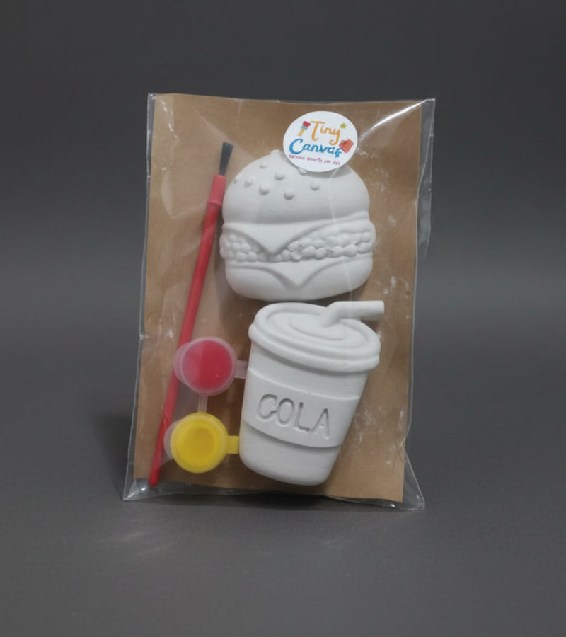 A packaged craft kit featuring a white unpainted ceramic or clay burger and a coffee cup, along with a paintbrush, small paint pots, sealed in a clear plastic bag on a brown surface.