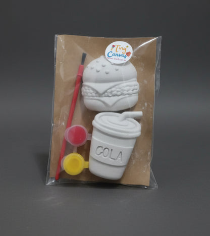 A packaged craft kit featuring a white unpainted ceramic or clay burger and a coffee cup, along with a paintbrush, small paint pots, sealed in a clear plastic bag on a brown surface.