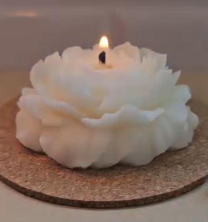 A white flower-shaped candle with a lit flame, resting on a round cork coaster on a light-colored surface.