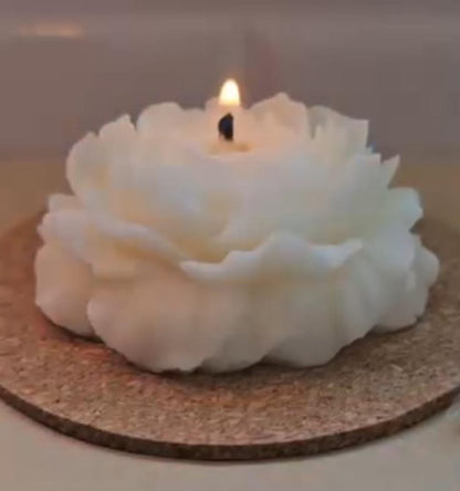 A white flower-shaped candle with a lit flame, resting on a round cork coaster on a light-colored surface.